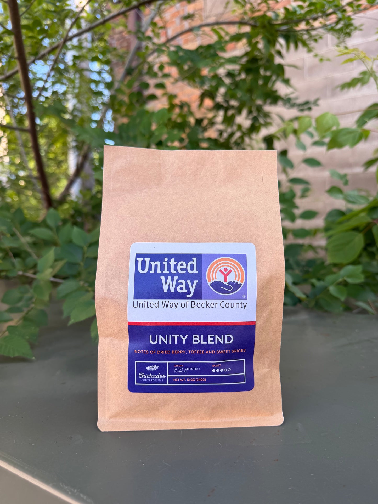 A brown bag of Unity Blend medium roast coffee from Chickadee Coffee Roasters sits outdoors among leafy plants and a brick wall. The label notes flavors of dried berry, toffee, and sweet spices.