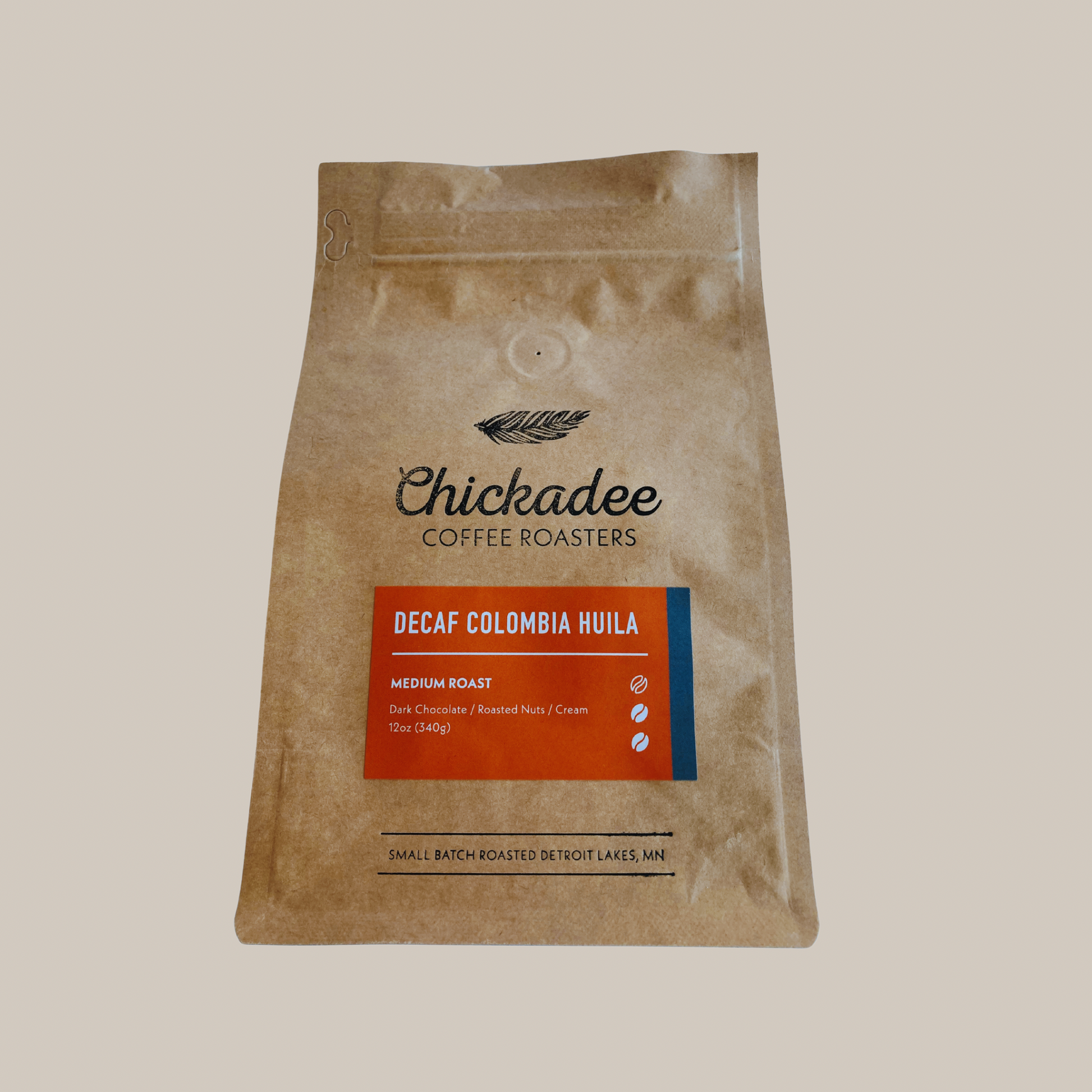A 12oz (340g) brown bag of Chickadee Coffee Roasters Decaf Colombian Huila, a medium roast with notes of dark chocolate, roasted nuts, and cream. This decaf coffee is made in Detroit Lakes, MN.