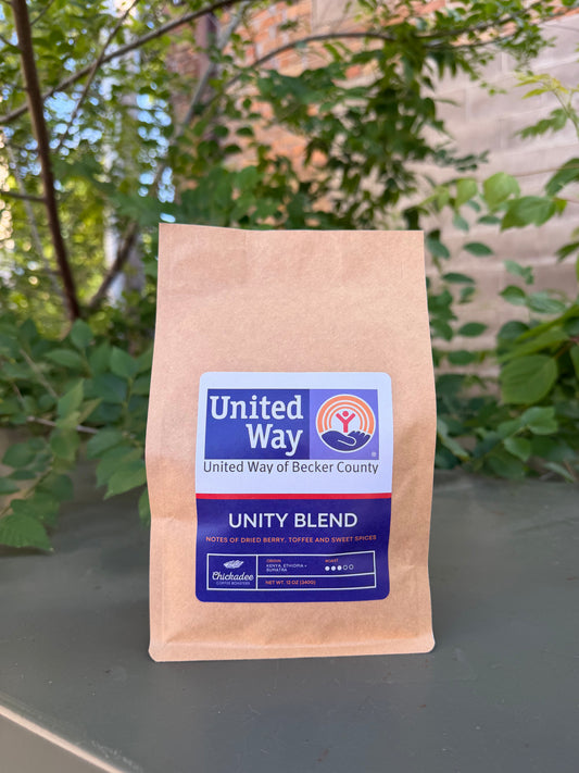 A brown bag of Unity Blend medium roast coffee from Chickadee Coffee Roasters sits outdoors among leafy plants and a brick wall. The label notes flavors of dried berry, toffee, and sweet spices.
