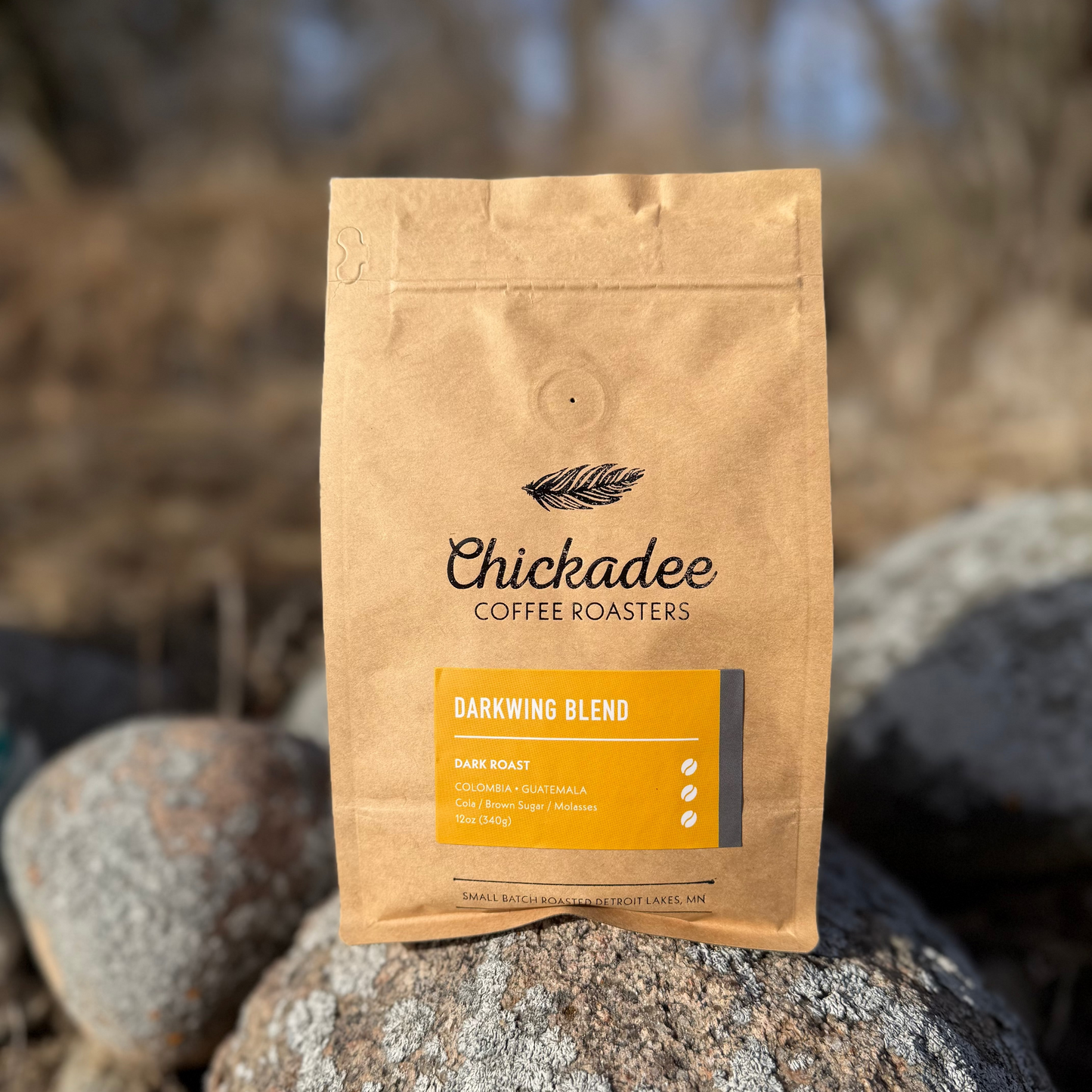 A brown bag of My Store Darkwing Blend sits outdoors on rocks. This bold dark roast coffee combines Colombian and Guatemalan beans with tasting notes of dark cherry, brown sugar, and molasses.