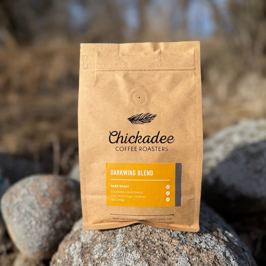 A brown bag of My Store Darkwing Blend sits outdoors on rocks. This bold dark roast coffee combines Colombian and Guatemalan beans with tasting notes of dark cherry, brown sugar, and molasses.