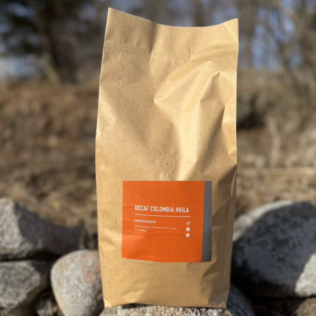 A brown paper bag labeled Decaf Colombian Huila from Chickadee Coffee Roasters rests on rocks outdoors, showcasing this Colombian decaf coffee with a medium roast and notes of red fruit and cocoa against a blurred natural backdrop.