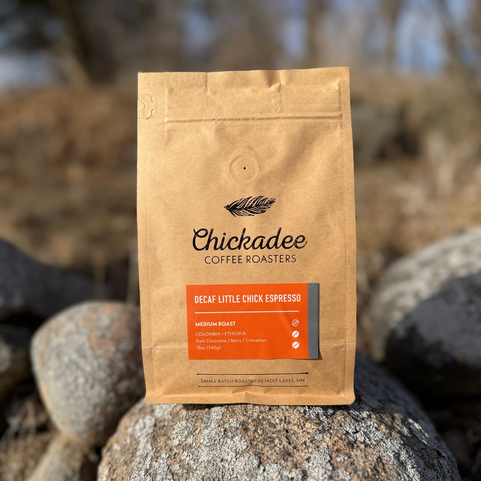 A brown bag of My Store Decaf Little Chick Espresso sits on rocks outdoors. The label highlights a medium roast with dark chocolate, cherry, and cinnamon notes, crafted using the sugarcane EA decaf process.