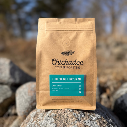 A brown bag of Chickadee Coffee Roasters’ Ethiopia Guji Kayon Mt coffee rests on a rock outdoors. This light roast offers blueberry, plum, and chocolate notes, set against a backdrop of blurred trees and rocks.