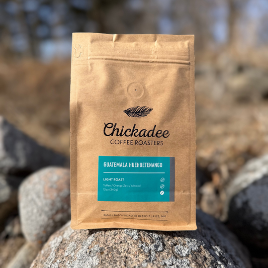 A brown bag of Chickadee Coffee Roasters’ Guatemala Huehuetenango sits on rocks outdoors, featuring tasting notes of toffee, orange zest, and almond.