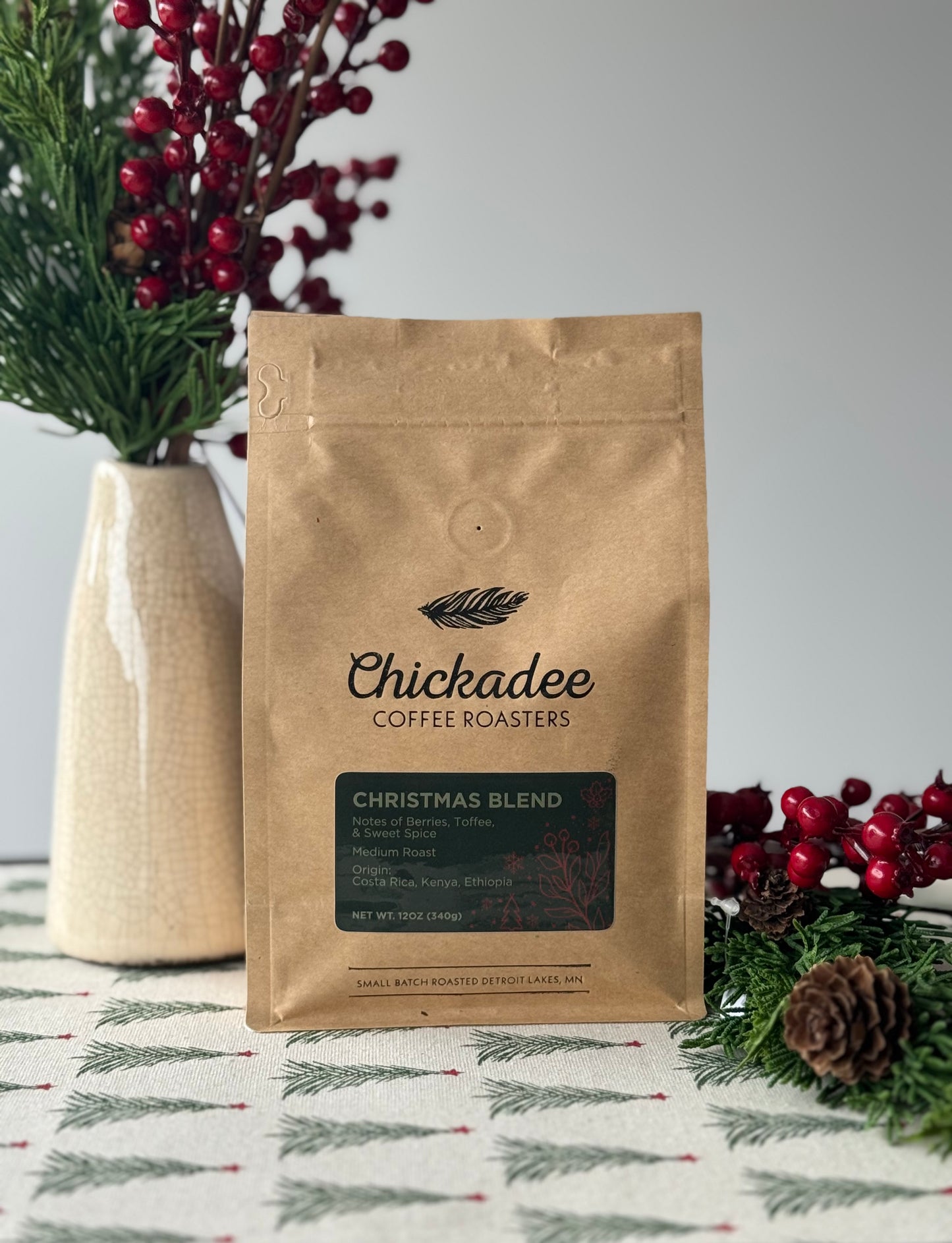 A brown bag of Chickadee Coffee Roasters Christmas Blend sits on a table with festive greenery, red berries, and a pine cone; in the background is a white vase filled with more greenery and berries.