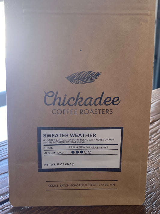 A brown 12 oz bag from Chickadee Coffee Roasters features the Sweater Weather Blend, with a feather logo. This medium roast combines Papua New Guinea and Kenyan beans for a bold flavor.