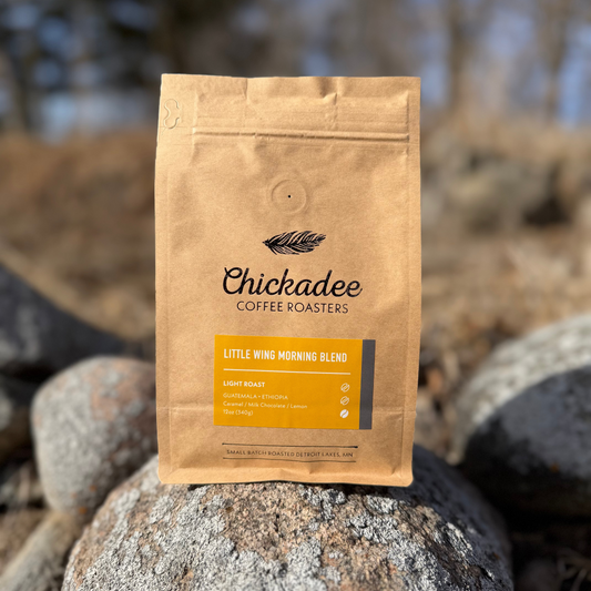 A brown paper bag of My Store's Little Wing Morning Blend sits on rocks outdoors. The label highlights this light roast, crafted from Ethiopia and Guatemala coffee, with chamomile and lemon notes.