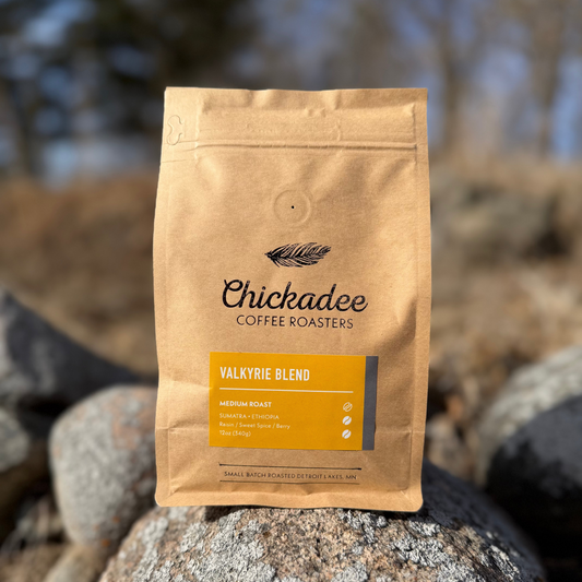A brown bag of Chickadee Coffee Roasters’ Valkyrie Blend coffee rests on rocks outdoors. This medium roast boasts flavors of tea, almonds, spice, and berry, with trees and sky softly blurred in the background.