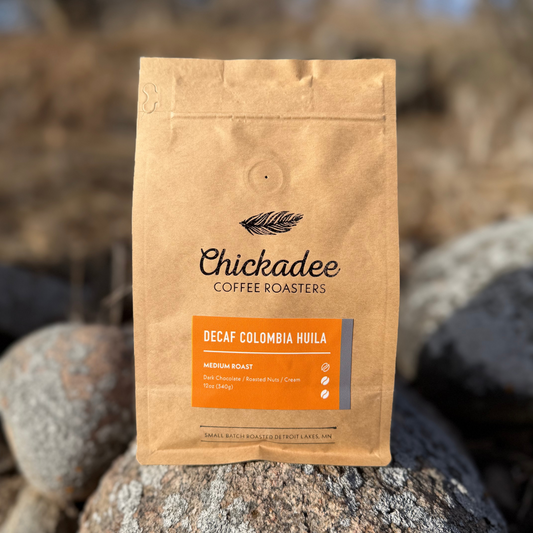 A brown bag of Chickadee Coffee Roasters Decaf Colombian Huila medium roast sits on rocks outdoors, with a label describing rich decaf coffee flavors including dark chocolate, roasted nuts, and cream.